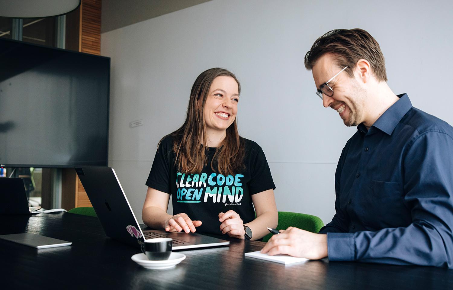 Zwei Personen sitzen an einem Laptop in einem Meetingraum, besprechen sich und lachen.