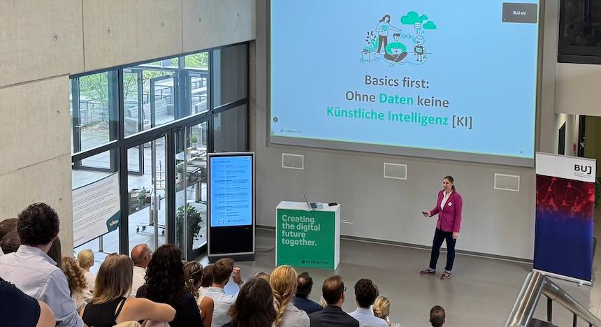 Vortrag im Rahmen des BUJ-Summit, auf Screen "Basics first: Ohne Daten keine KI", davor Vortragende, im Vordergrund Plenum