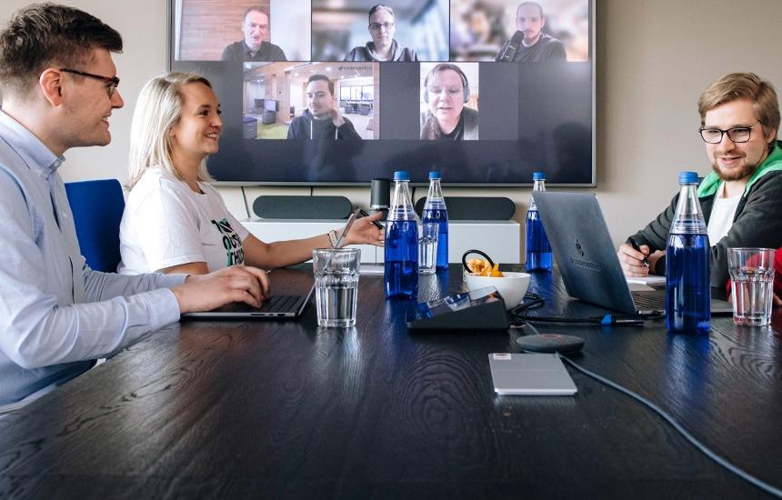 Projektbesprechung: drei Personen in einem Meetingraum, 5 weitere per Videokonferenz auf dem Bildschirm an der Wand dabei