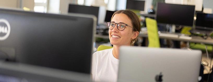 Eine Frau sitzt an einem Arbeitsplatz, schaut auf ihren Monitor und lächelt beim Arbeiten