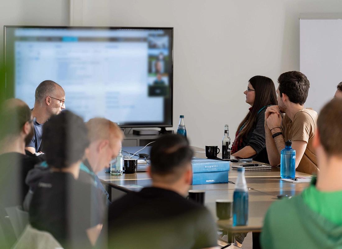 Projektgruppe sitzt in einem Meetingraum und weitere Personen per Videokonferenz sprechen über ein geteiltes Dokument
