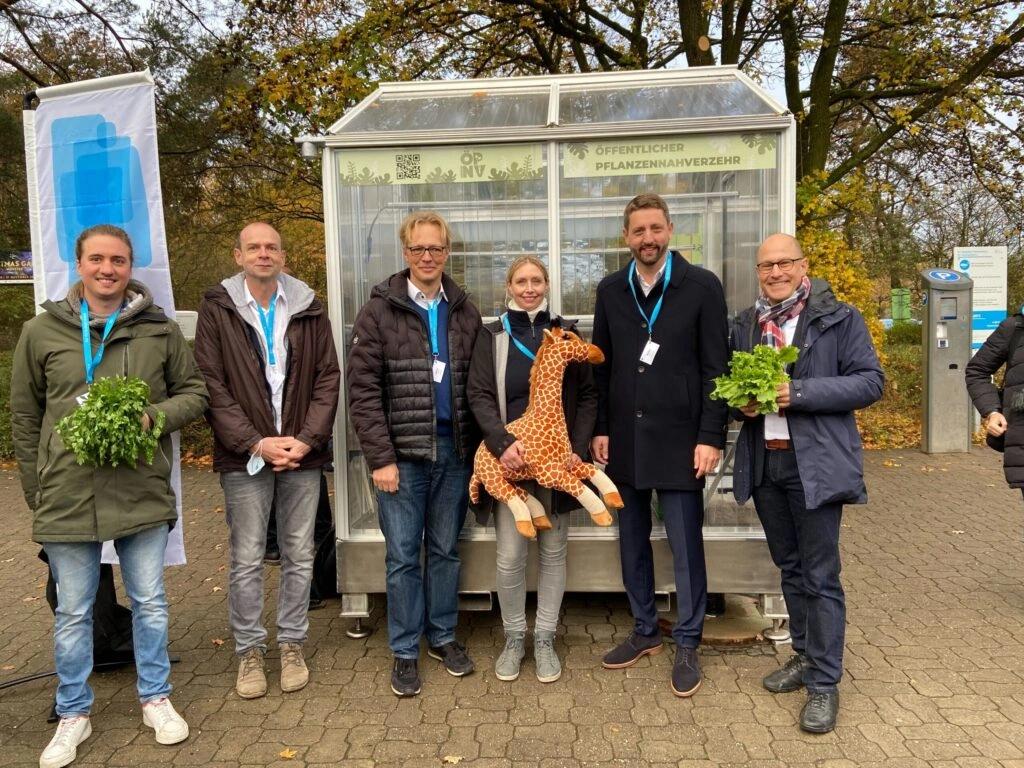5 Personen, die vor einem Gewächshaus stehen, die äußersten haben Salat und die in der Mitte eine Plüschgiraffe in der Hand.