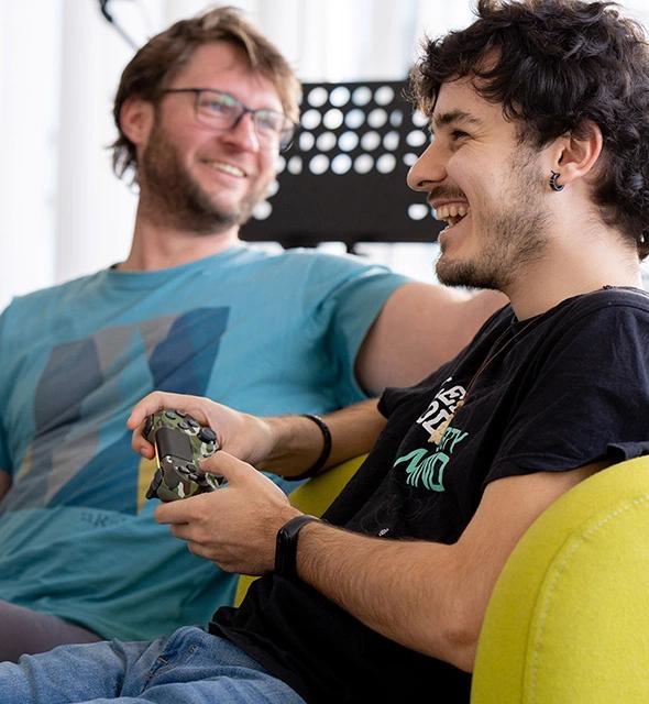 Zwei Männer sitzen lachend auf einem Sofa, einer hält einen Controller für Videospiele in der Hand
