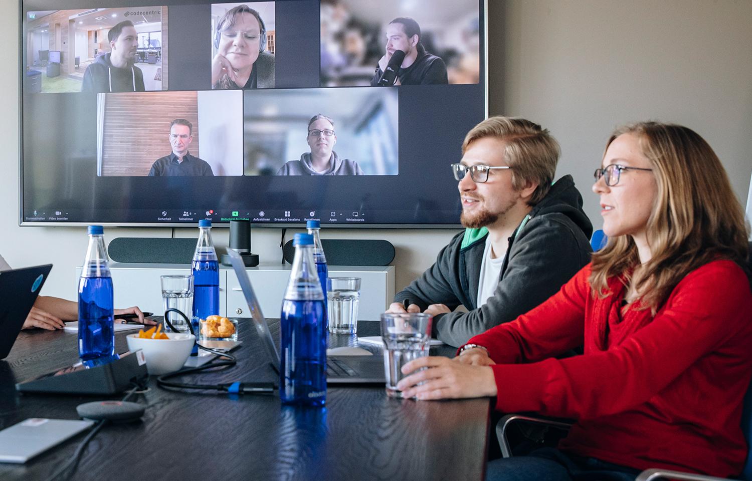 Projektbesprechung: drei Personen in einem Meetingraum, 5 weitere per Videokonferenz auf dem Bildschirm an der Wand dabei